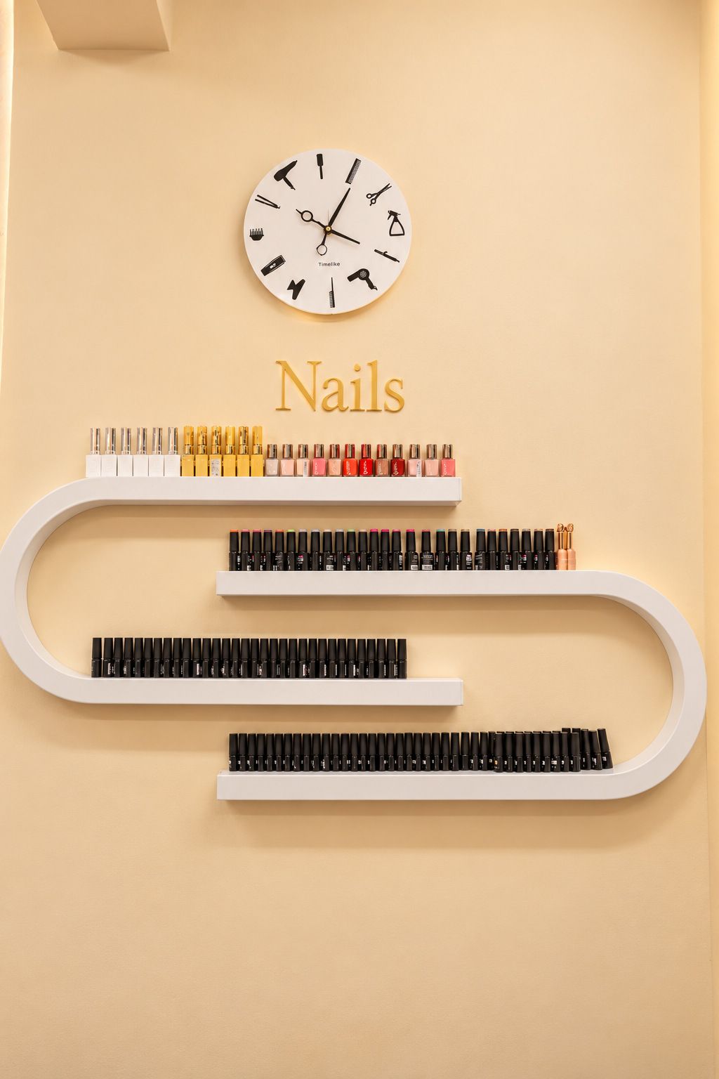 Gel polishes on curved white shelves under gold Nails lettering and a wall clock
